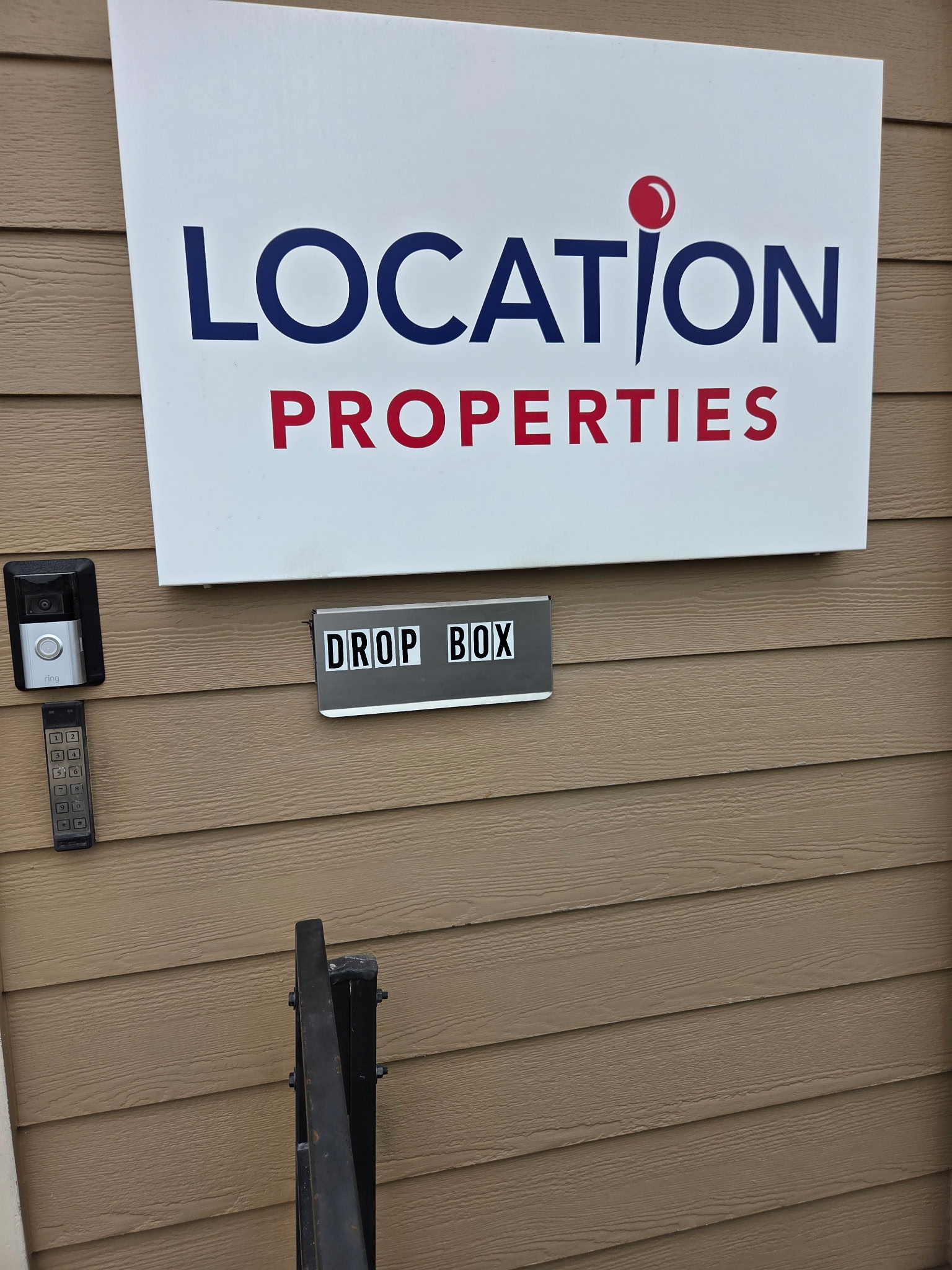 The metal DROP BOX slot mounted on the wall by the Location Properties office entrance at 3300 Clinton Parkway Court, Suite 110, below the Location Properties sign and beside the Ring doorbell camera